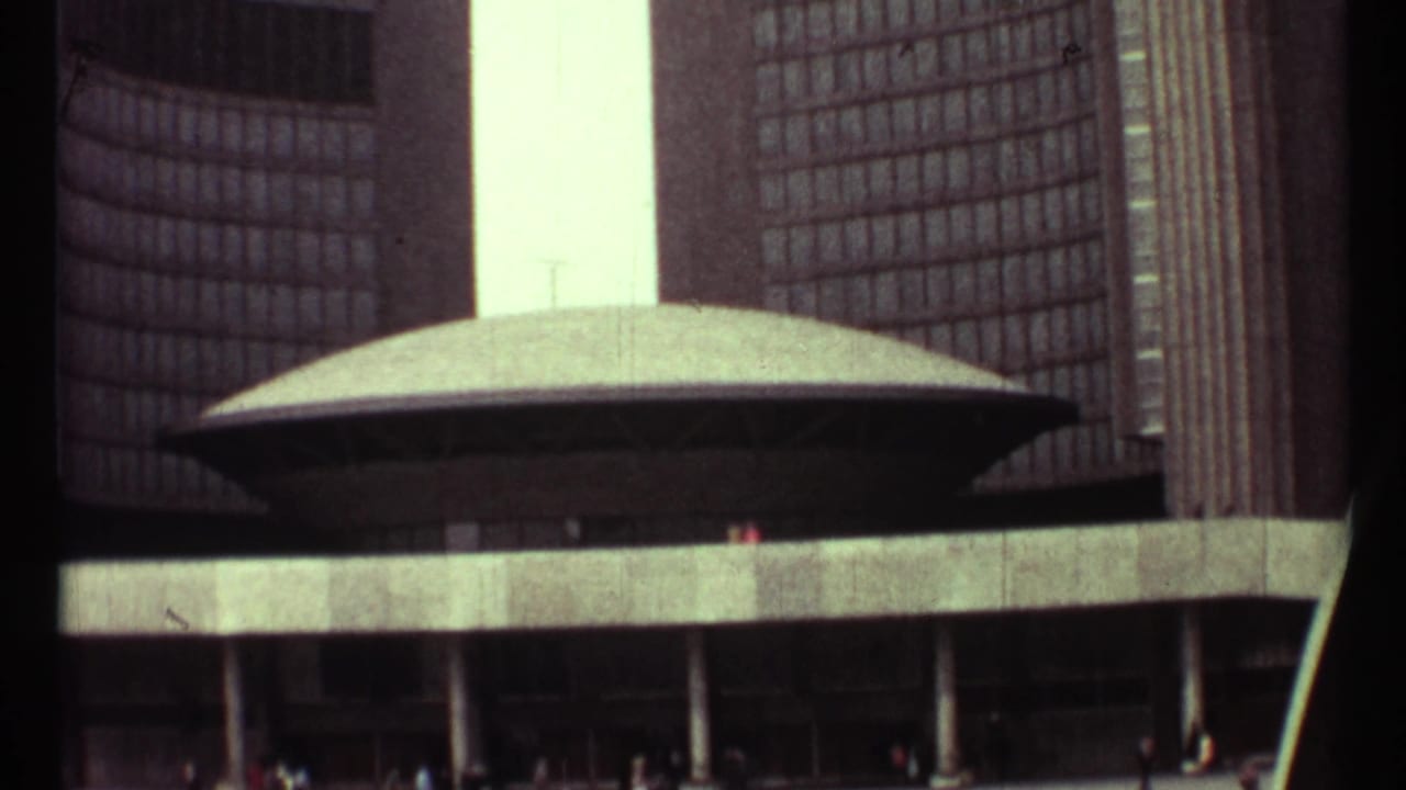 1967 8mm view of Toronto City Hall with curved towers, oval council chamber roof, concrete plaza edge, and small pedestrians below.