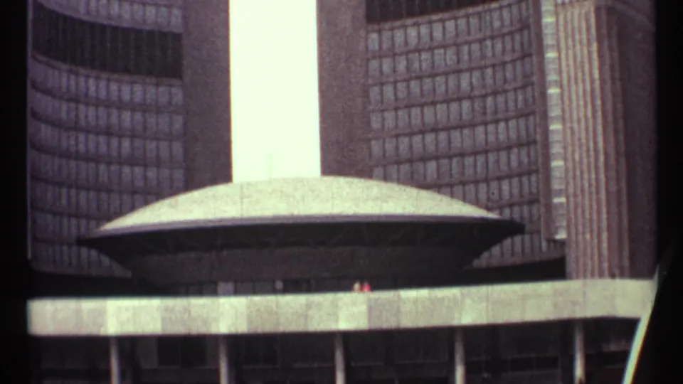 A closer 1967 8mm frame shows Toronto City Hall's oval council chamber, gridded tower windows, concrete ledge, columns, and distant plaza visitors.