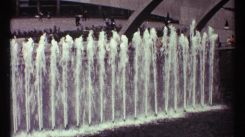 1967 Toronto 8mm frame showing a public fountain with multiple water jets and distant people walking near modernist buildings.