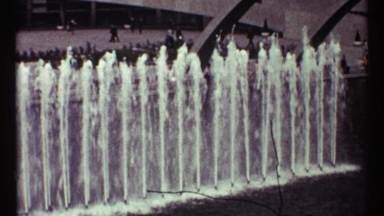 Tall fountain jets rise in front of a modern building with distant pedestrians above the pool.