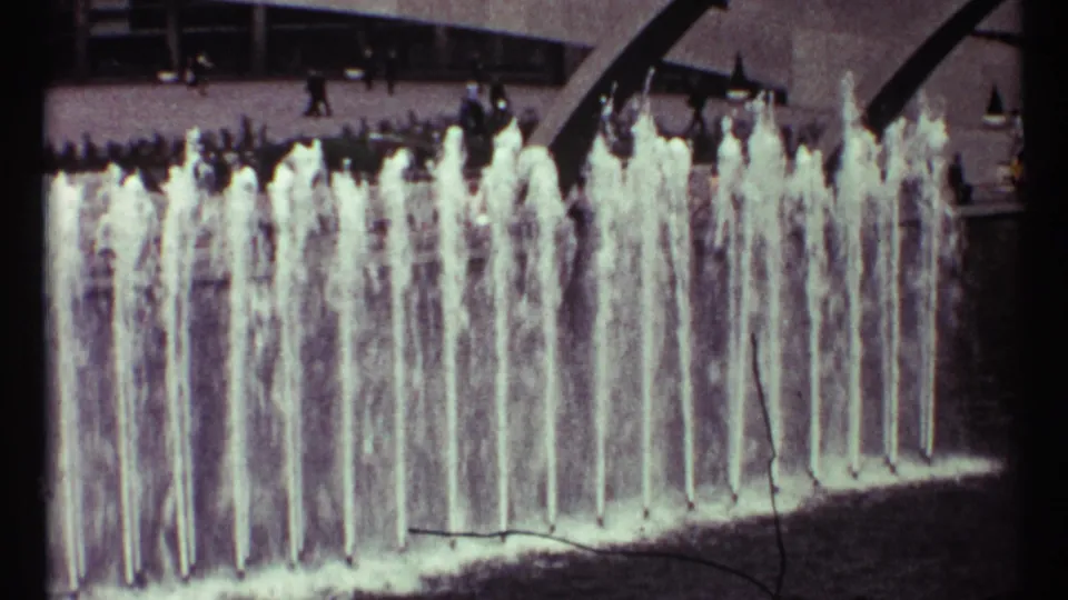 1967 Toronto 8mm frame showing vertical water jets in a public fountain beside a modern building and passing pedestrians.