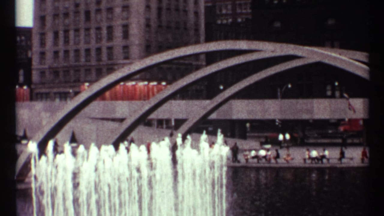 1967 Toronto home-movie frame showing fountain jets, sweeping concrete arches, pedestrians, vehicles, and tall downtown buildings.