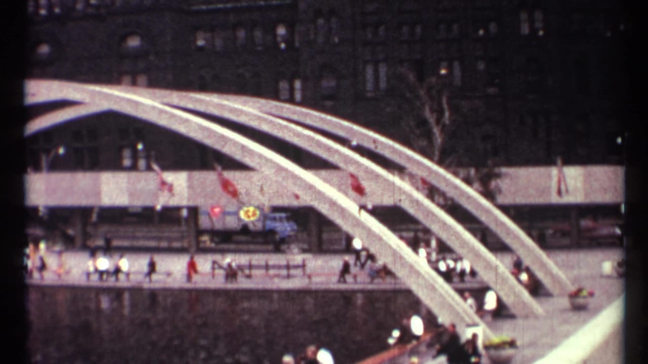 White arched structure over a plaza with pedestrians, flags, and dark buildings behind it.