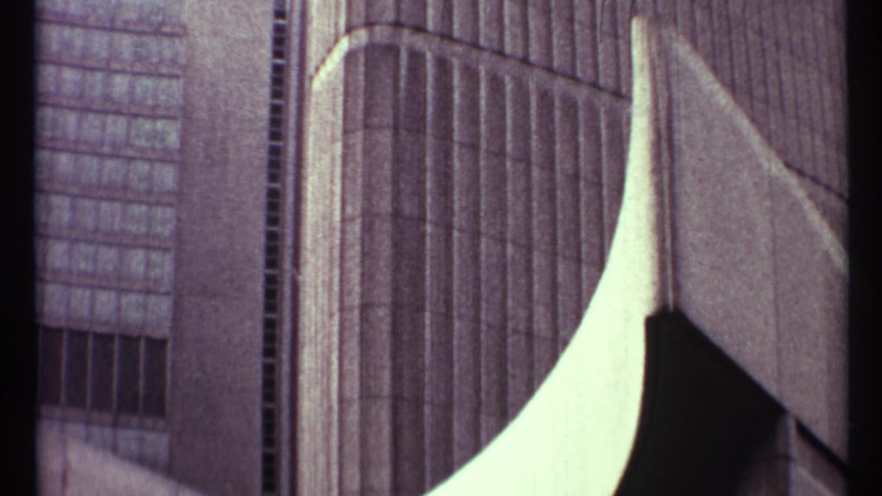 Soft 8mm frame from the 1967 Toronto clip showing tall urban building facades and a pale curved architectural or street-side form in the foreground.