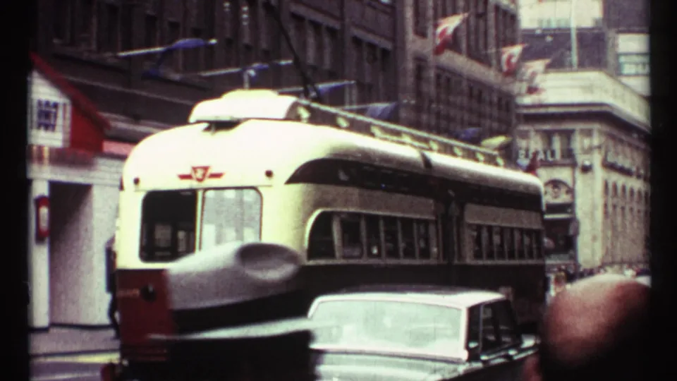 1967 Toronto street scene with a red-and-white electric streetcar under overhead wires, a car in front, storefronts, and flags above the sidewalk.