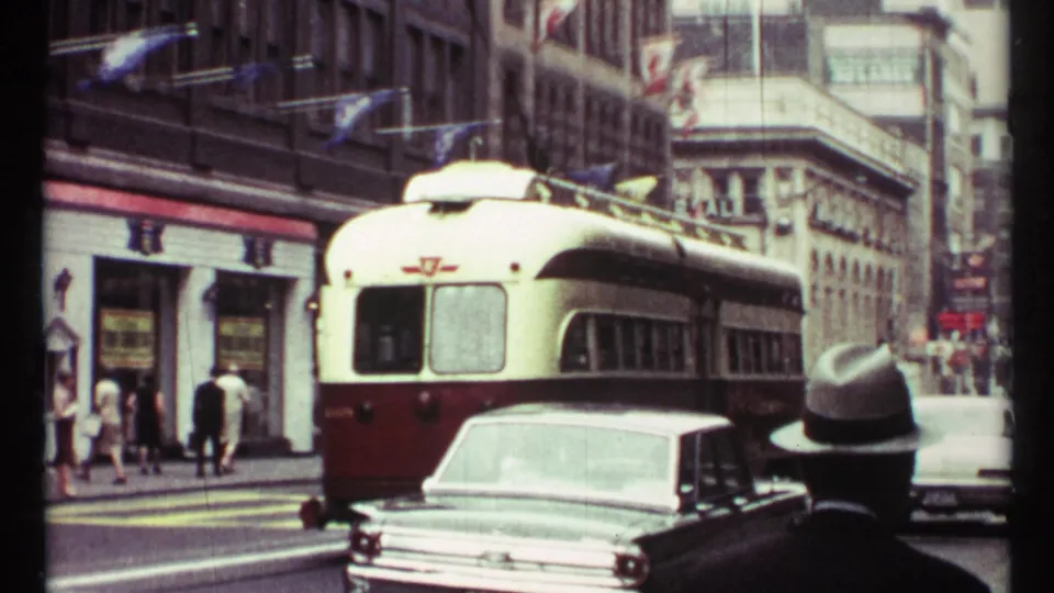 1967 Toronto street view showing a red-and-white streetcar behind a mid-century car, pedestrians on the sidewalk, storefronts, and flags along the buildings.