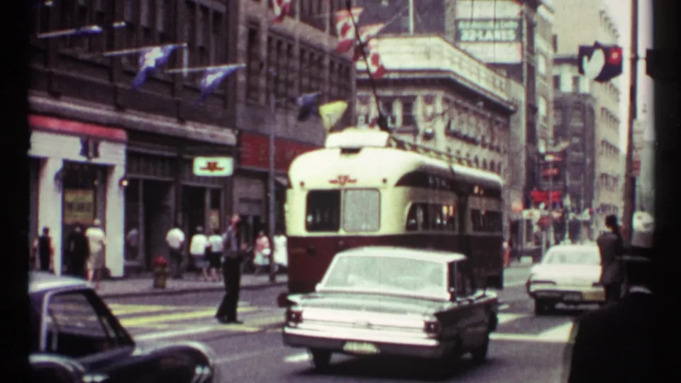 1967 Toronto frame showing a red-and-white electric streetcar in traffic, a light-colored car ahead, pedestrians on both sidewalks, overhead wires, and flags on building fronts.
