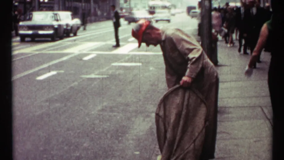 Toronto 1967 street frame showing a sidewalk cleaner in a red cap or bandana bending over a large brush near pedestrians and cars.