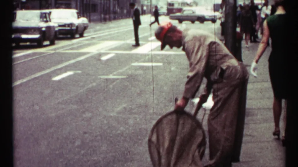 Toronto 1967 home-movie frame with a worker using a large street brush near the curb as pedestrians and cars occupy the background.