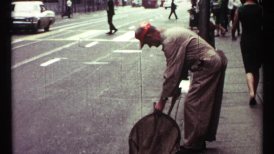 Toronto 1967 street frame showing a sidewalk cleaner holding a large brush near the curb, with passing pedestrians and traffic behind.