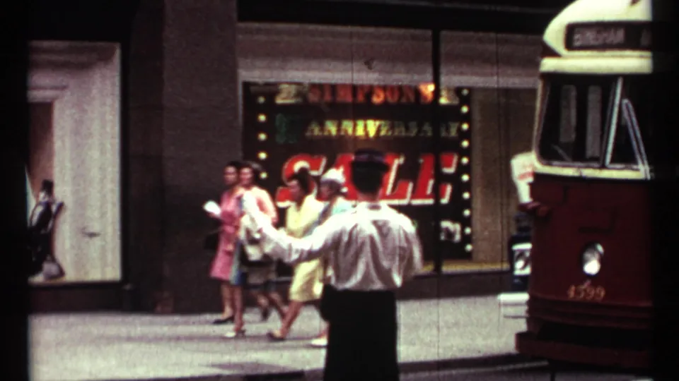 Traffic director in white shirt and gloves manages pedestrians near a Simpson's sale window as a red-and-cream vehicle enters frame.