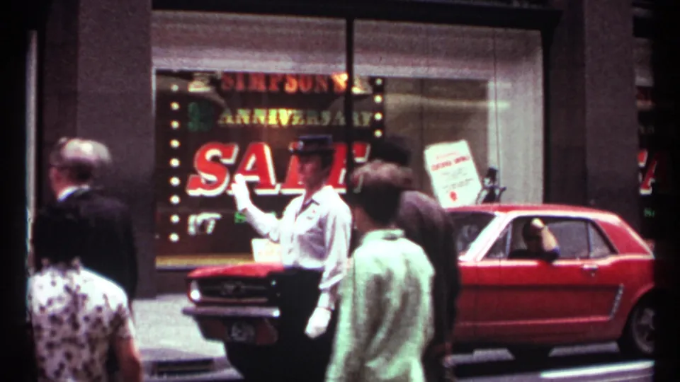 A man in a white uniform and cap gestures near a parked red Mustang outside a Simpson's anniversary sale window in Toronto.