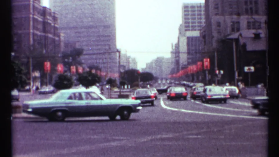 A light blue vintage sedan crosses a 1967 Toronto street as other cars travel past buildings and red banners.