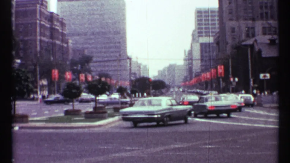 1967 Toronto street traffic with several vintage sedans moving along a broad road beneath rows of red banners.