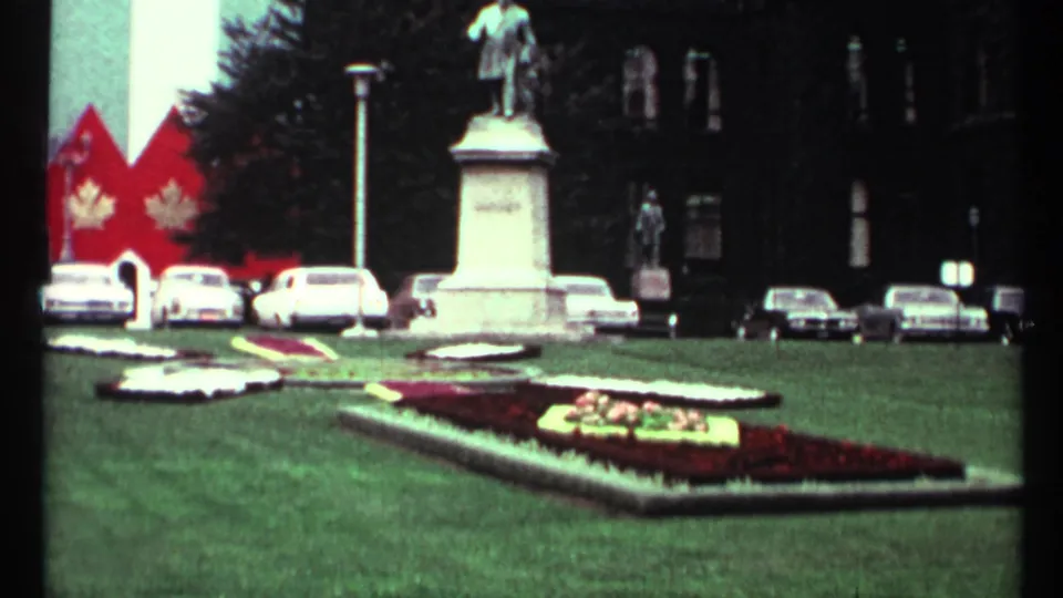 1967 Toronto home-movie view across formal flower beds toward a statue pedestal, parked cars, trees, and red maple-leaf banners.