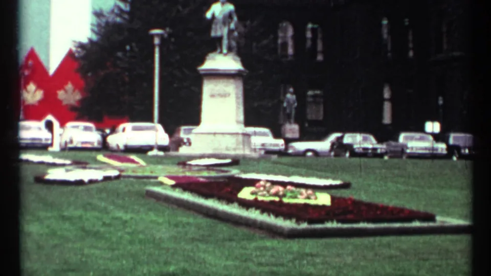 1967 Toronto civic grounds with a statue, patterned flower beds, parked 1960s cars, red maple-leaf banners, and a dark building backdrop.