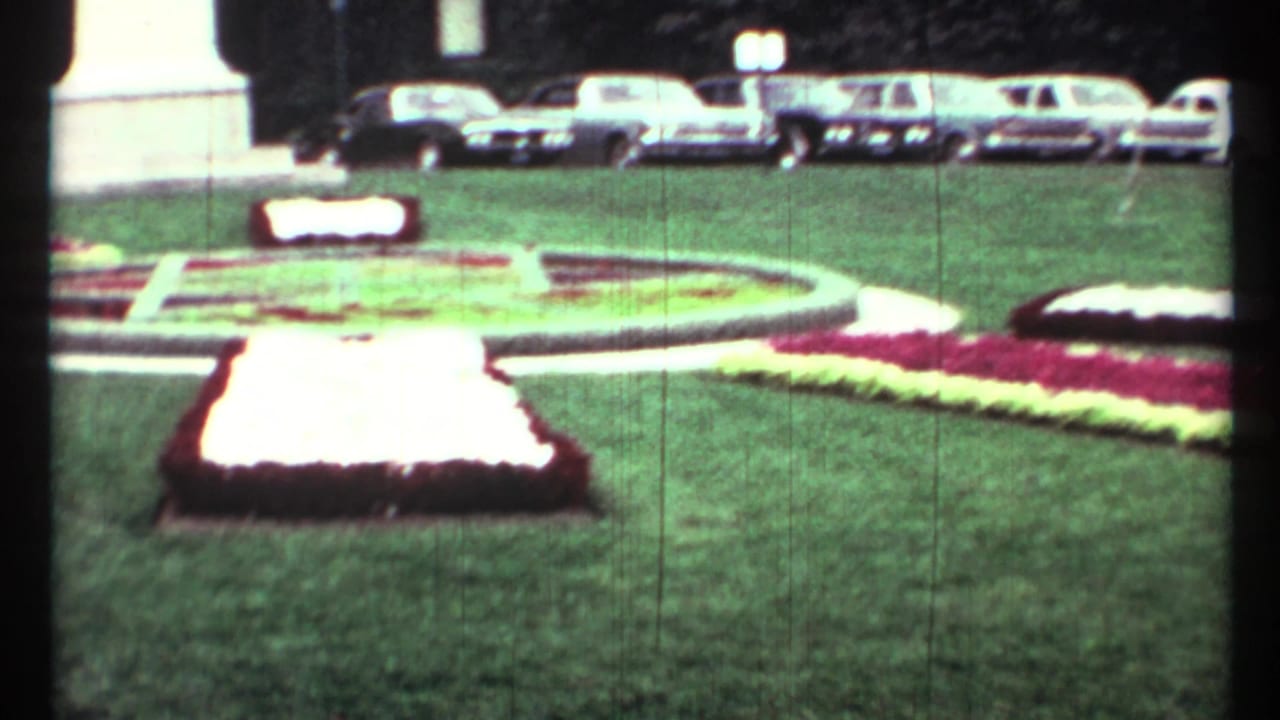 Flower beds and lawn in a civic square with parked classic cars behind a monument base.