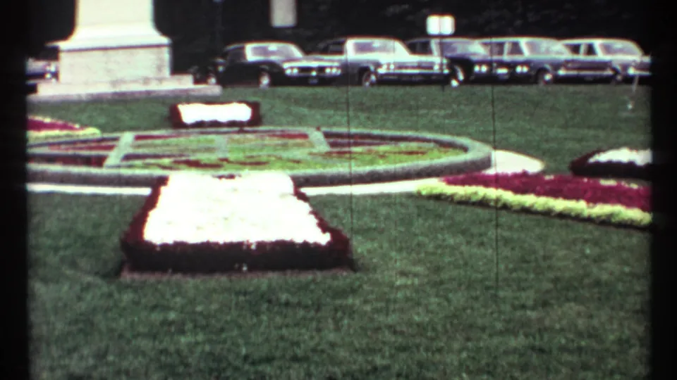 Toronto civic-square view with clipped lawn, formal floral planting, monument base, and 1960s parked cars in the background.