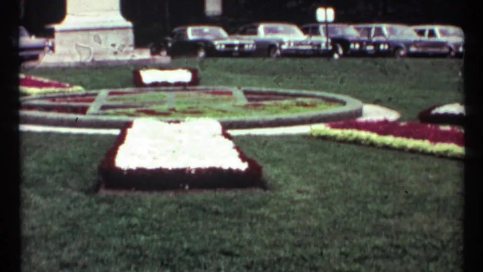 1967 Toronto frame centered on a bright rectangular flower bed, green lawn, monument base, and parked automobiles.