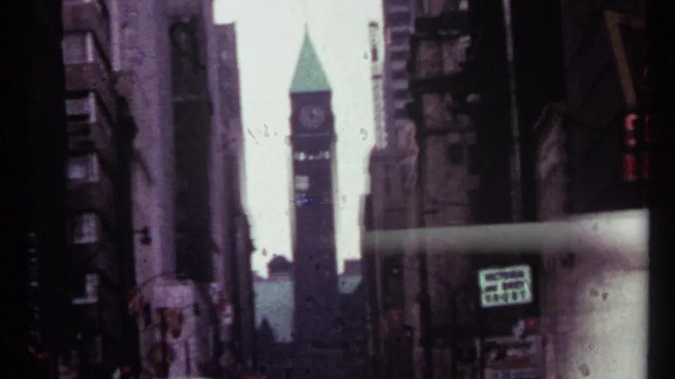 1967 Toronto street view looking between tall buildings toward a clock tower with a green spire and a visible commercial sign.