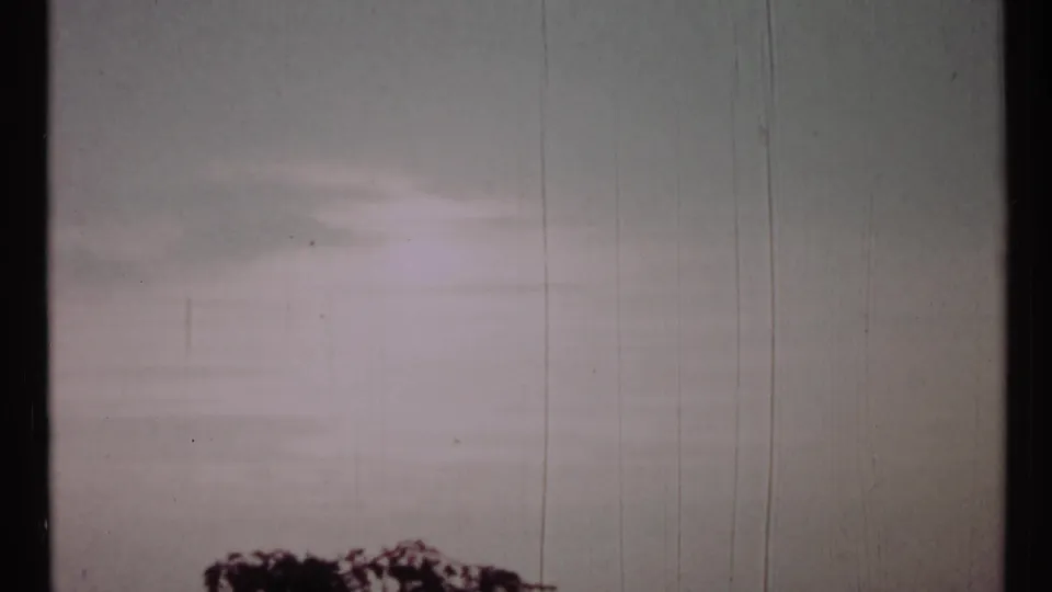 1967 Toronto 8mm frame with pale sky, faint flare, vertical film scratches, and dark treetop shapes along the lower edge.