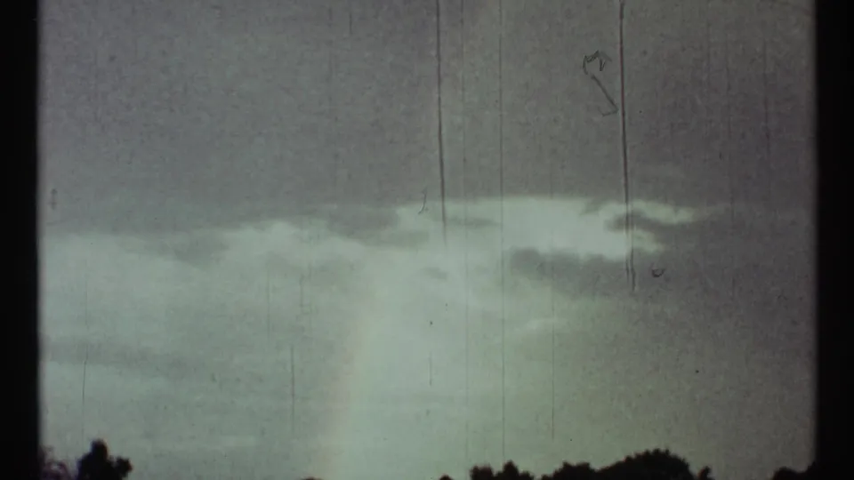 Faint rainbow near the horizon in faded 1967 8mm footage, with dark trees and open land below the cloud cover.
