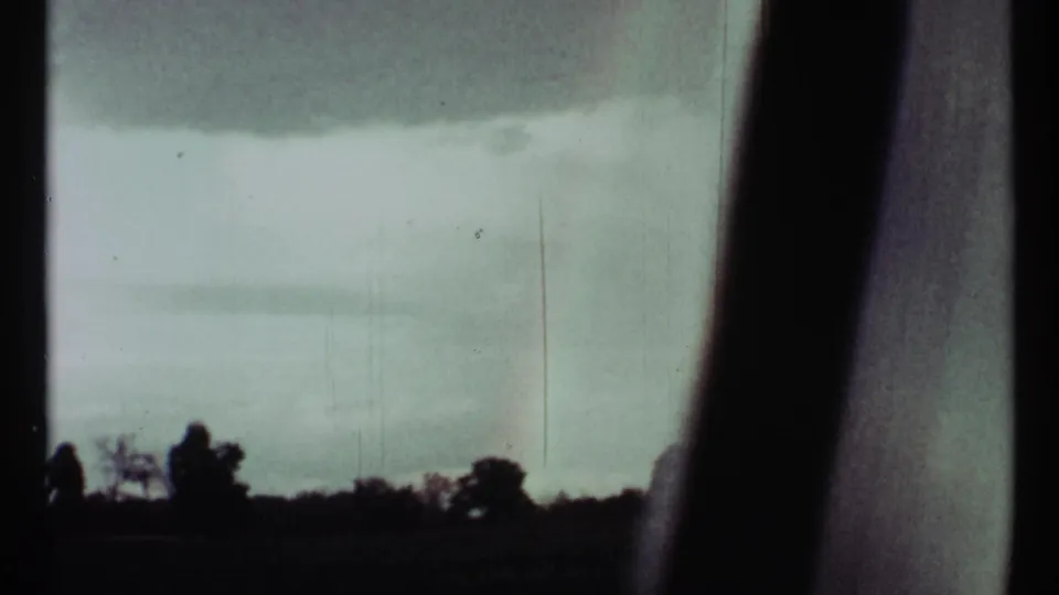 Faded 8mm view of a cloudy rural horizon and faint rainbow, with heavy dark film edge visible along the right side.