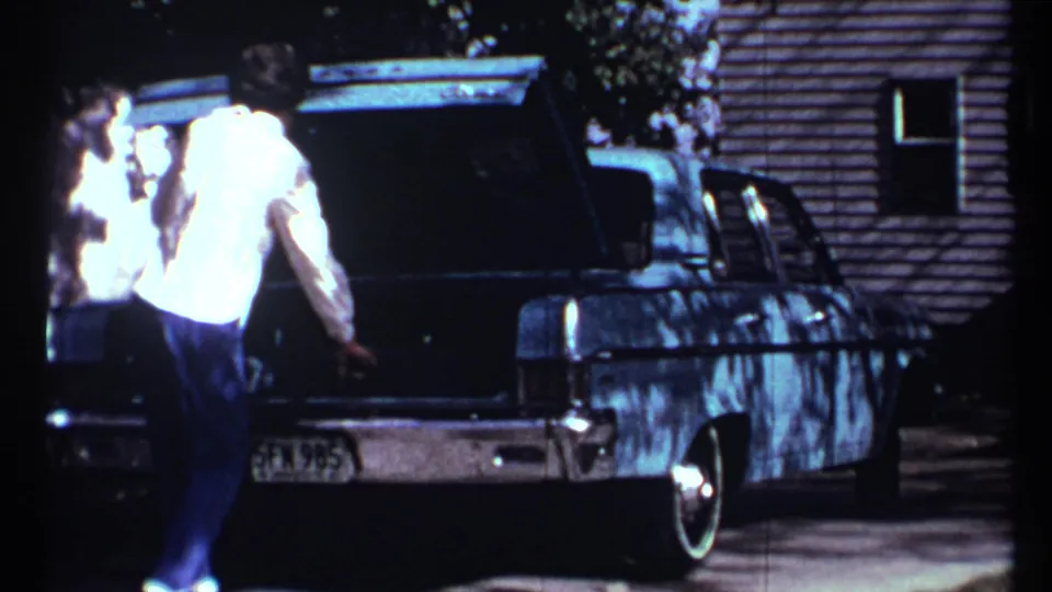 A man in a light shirt stands by the open trunk of a classic sedan in a Toronto home-movie driveway scene.