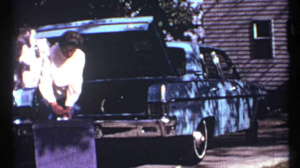 A man bends toward luggage beside an open sedan trunk as another person stands at the edge of the frame in 1967 Toronto footage.