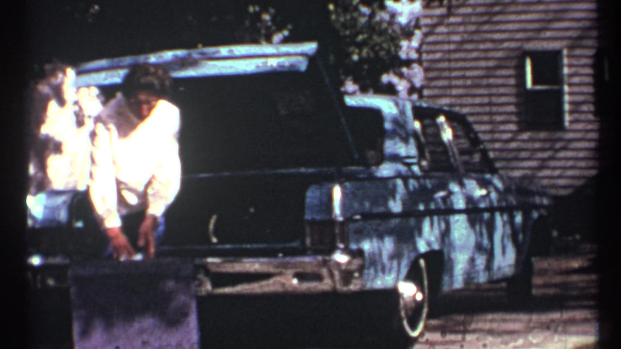 In 1967 Toronto home-movie footage, a woman in a light sweater bends near a dark sedan with its trunk open as a white cat stands nearby.