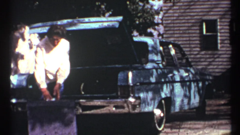 In 1967 Toronto home-movie footage, a woman in a light sweater bends near a dark sedan with its trunk open as a white cat stands nearby.