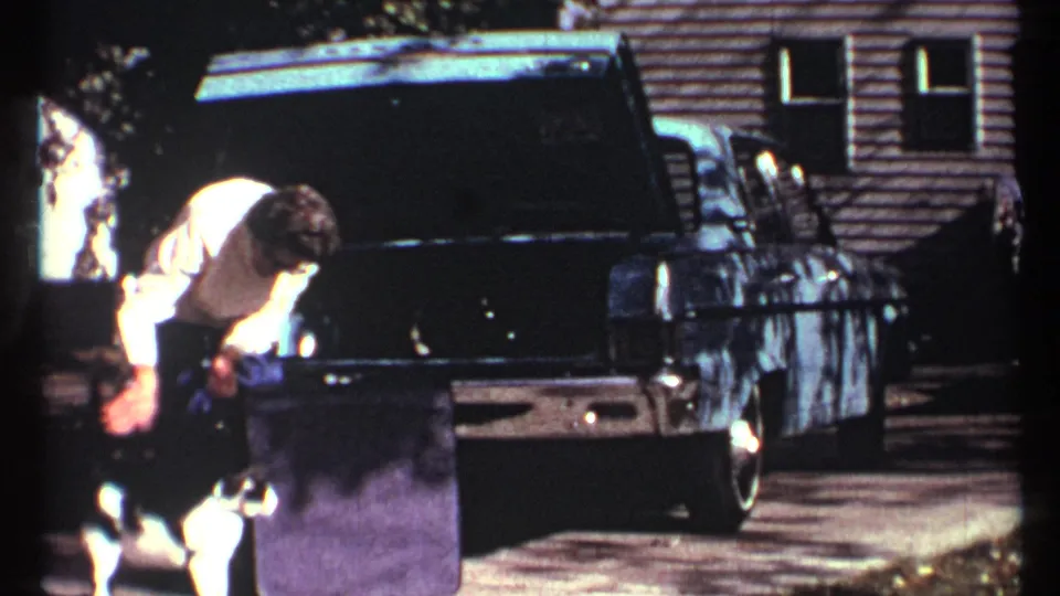 The woman leans toward the white cat beside the open trunk of a dark sedan, with a house exterior visible behind them.