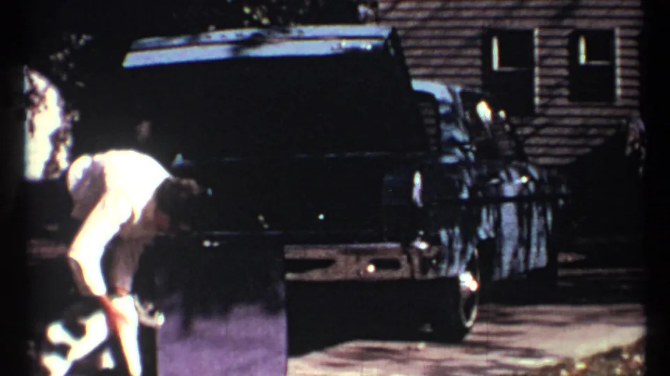 A woman bends low beside the white cat at the rear of the dark sedan, with strong sunlight and soft 8mm detail across the frame.
