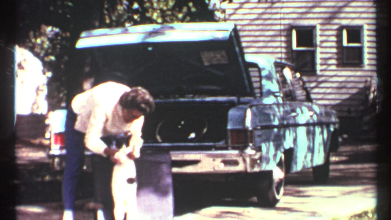 In 1967 Toronto home-movie footage, a man bends near an open blue car trunk while holding a black-and-white cat outdoors.
