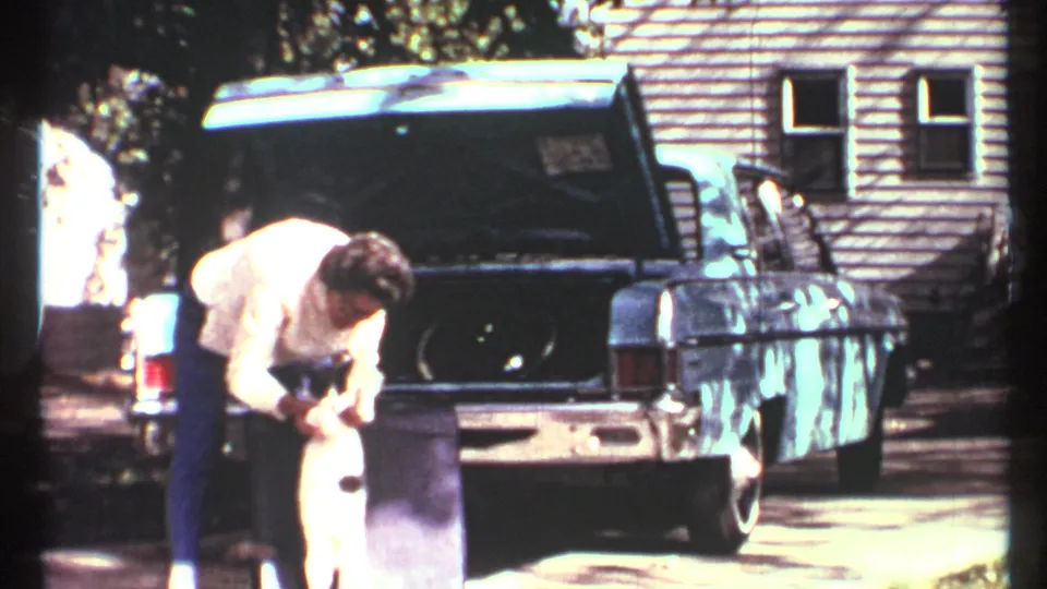 In 1967 Toronto home-movie footage, a man bends near an open blue car trunk while holding a black-and-white cat outdoors.