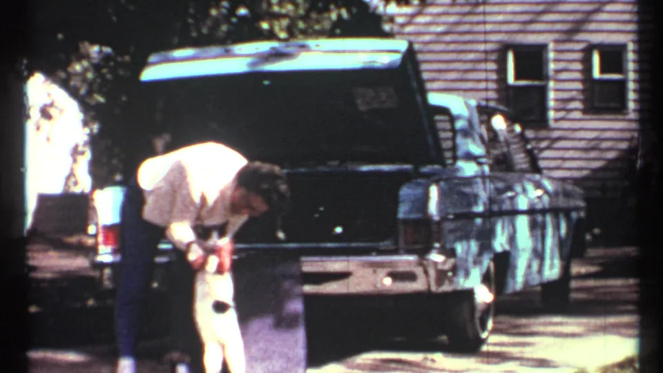 The man remains bent toward the black-and-white cat, with the blue car trunk open behind him in the Toronto residential scene.