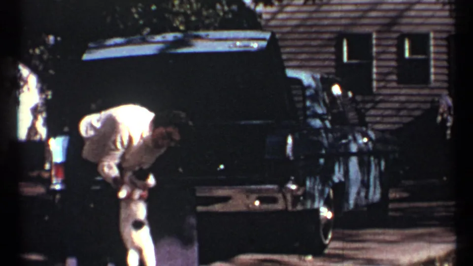 At the end of the clip, the man bends beside the cat while the open blue car trunk dominates the residential background.