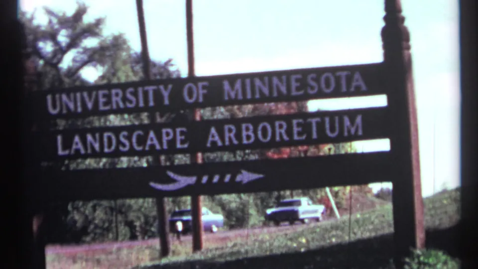1967 8mm frame showing a wooden University of Minnesota Landscape Arboretum sign with white lettering, arrow, trees, grass, and cars beyond it.