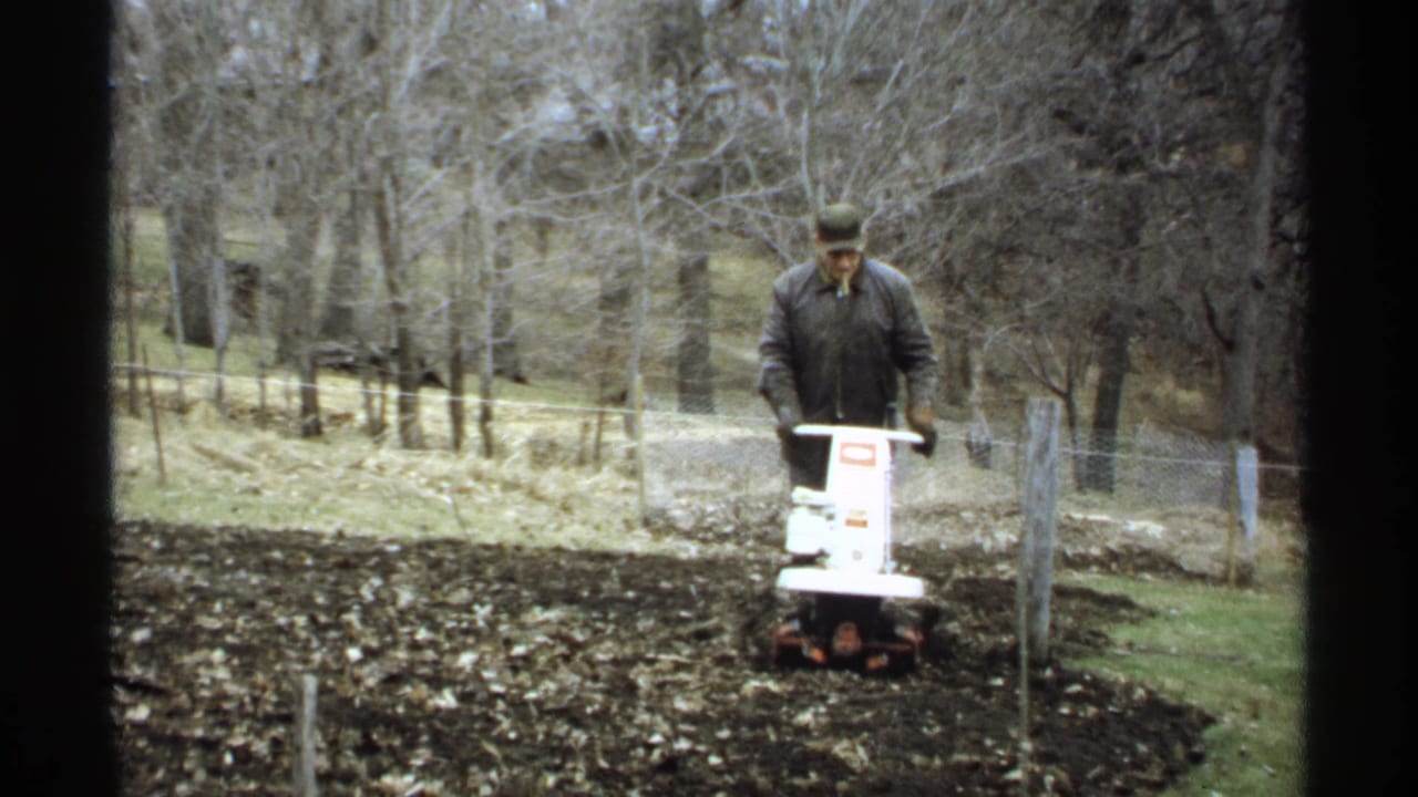 Man operating a white garden tiller in a rural plot with bare trees and fencing behind him.