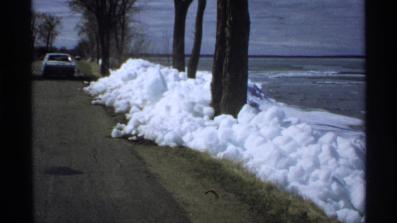 Snowbanks line a quiet road beside a lake with bare trees and a parked car.