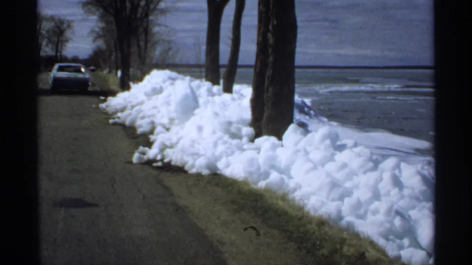 A quiet Minnesota lakeside road appears beside melting snowbanks, bare trees, and a parked car in early-1970s 8mm footage.