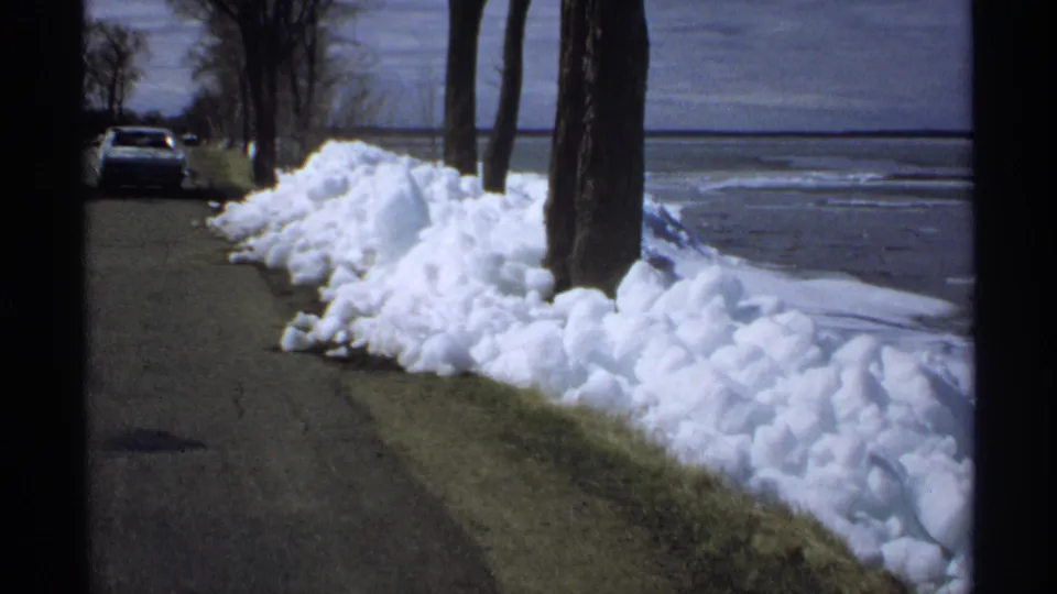 The camera looks along a lakeside road in Minnesota, with melting snow piled beside trees and a parked car ahead.