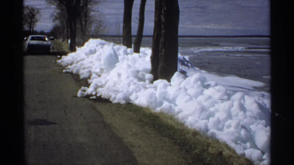 A final 8mm frame shows the same quiet Minnesota lakeside road with snowbanks, bare trees, and a parked car near the water.