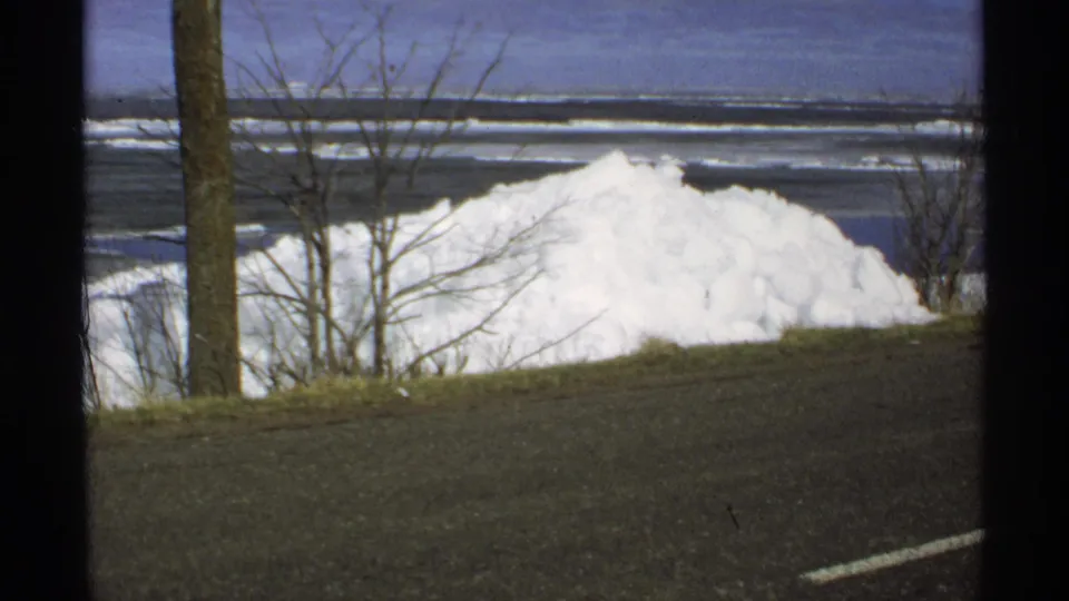 1970 Minnesota roadside view with a large snowbank, bare trees, pavement, and a frozen lake or icy shoreline beyond.