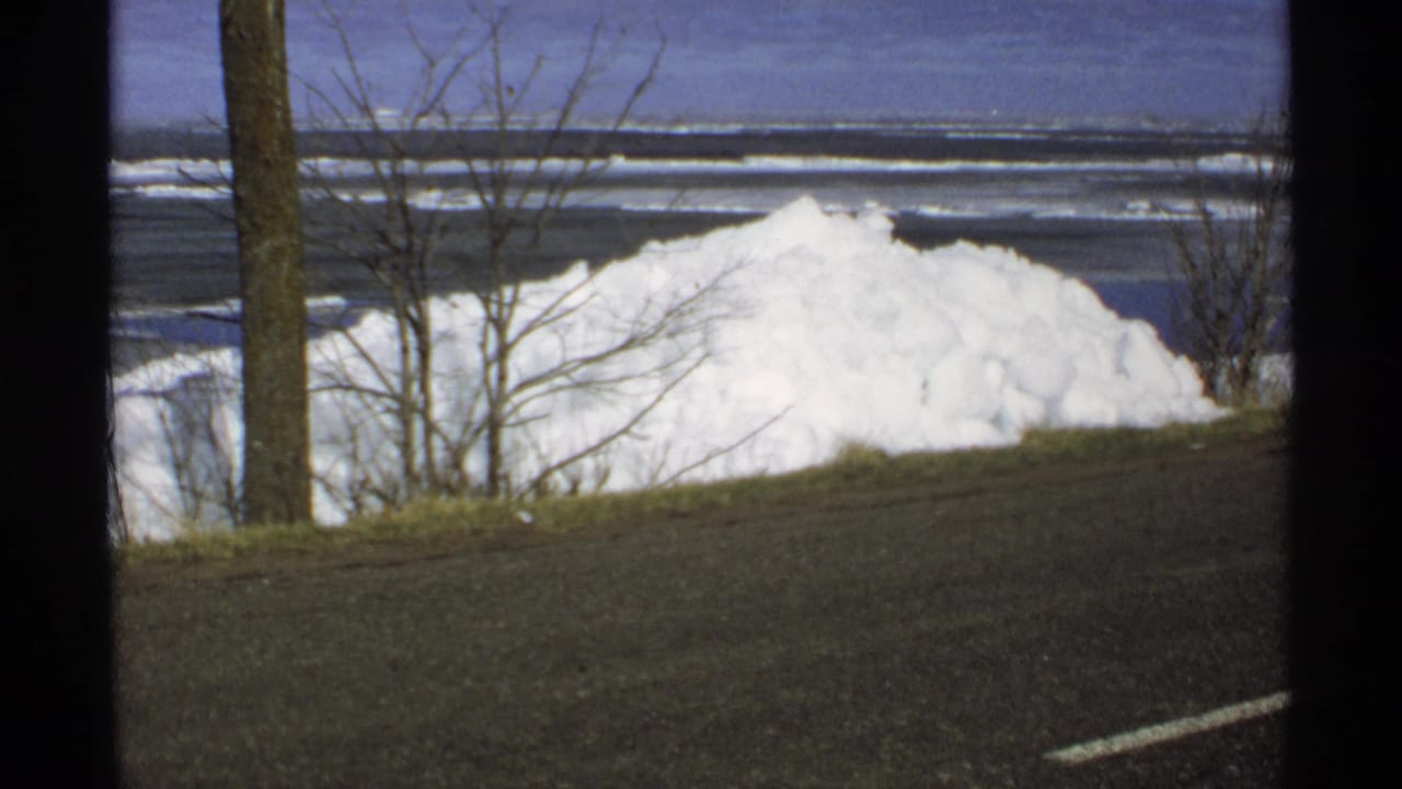 Large snowbank beside a road with bare trees and a frozen lake beyond.