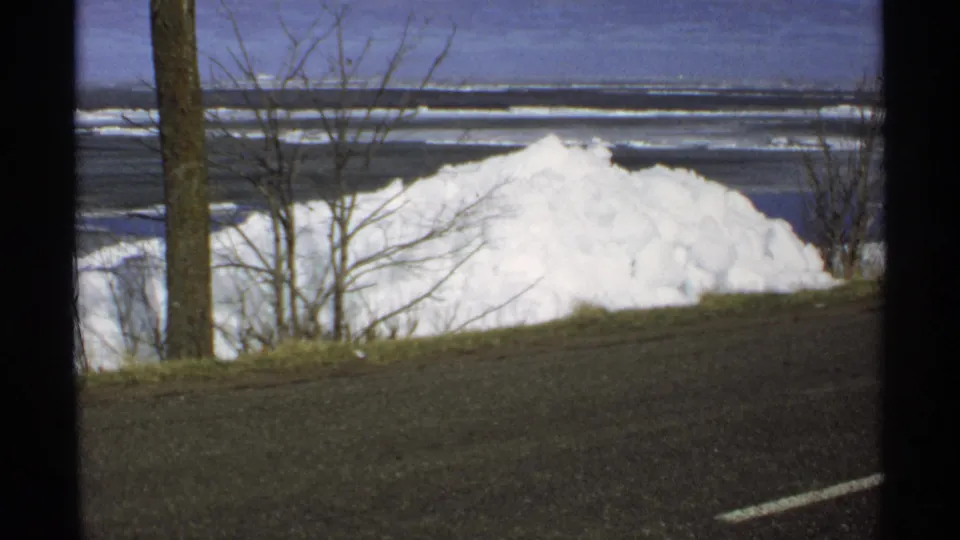 1970 Minnesota 8mm frame showing a large roadside snowbank, bare trees, a dark road edge, and frozen lake water in the background.