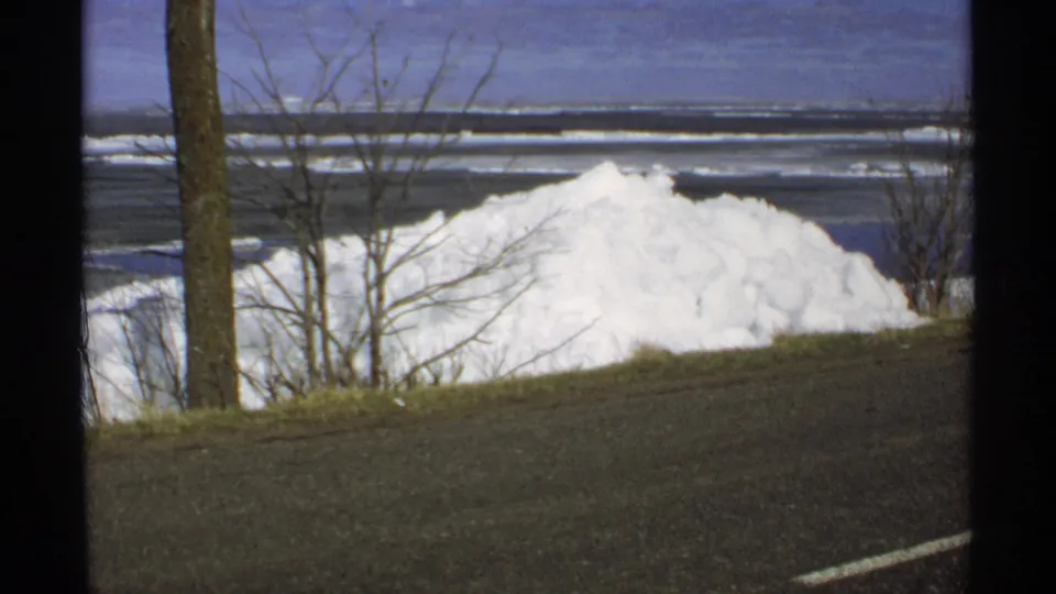 1970 Minnesota roadside view with a piled snowbank, bare branches, tree trunks, and a frozen lake beyond the slope.