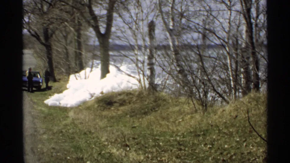 1970 Minnesota home-movie frame showing a lakeside road, melting snowbanks, bare trees, a blue car, and two distant people near the roadside.