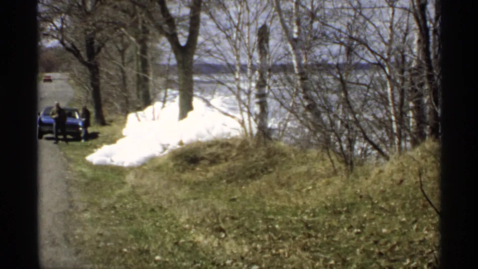 1970 Minnesota roadside frame with a blue car and distant person on a narrow road, bordered by bare trees, snowbanks, grass, and frozen lake water.