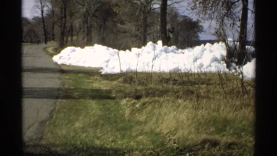 1970 Minnesota 8mm frame with a narrow wooded road, bright snowbanks, spring grass, bare trees, and water in the distance.